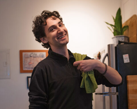 Nick Van Deren poses in his kitchen, wearing a black sweater with a green kitchen towel over his shoulder
