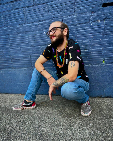 Alex Titus poses smiling next to a dark blue wall, wearing a colorful shirt and jeans.