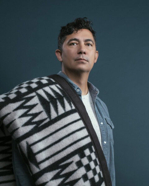 dramatic portrait of louie gong in an unbuttoned blue dress shirt with a Native-inspired blanket draped over his shoulder