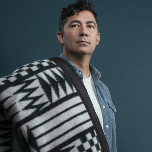 dramatic portrait of louie gong in an unbuttoned blue dress shirt with a Native-inspired blanket draped over his shoulder