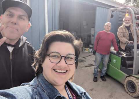 Meg is taking a selfie at the Loon Lake Food Bank with Ernest on the left, Terry on the forklift, and Henry near the forklift. Terry has a box of Northwest Harvest food on the forks.