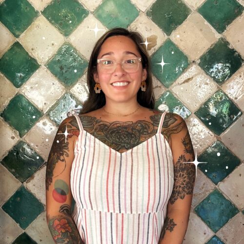 Tattooed smiling Marisol in sleeveless dress against tile wall