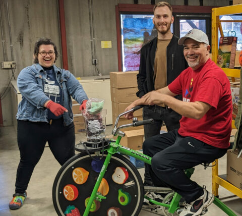 Scott is on the electric bike that they use to provide food demonstrations for children. Fruit is in the blender. Northwest Harvest staff are next to Scott.